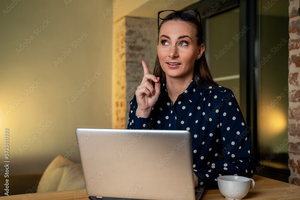 Fototapeta premium A woman sitting at her workplace and looking at a laptop screen, raising her index finger, feels happy and proud, making the right creative decision. A self-employed woman came up with a great