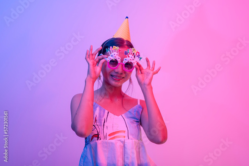 A young and cheerful female celebrant wears cute novelty birthday glasses. An asian lady partying. Plain background lit with blue and pink neon colors. © Mdv Edwards