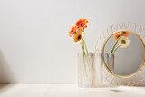 white glass vase with orange and beige flowers stand on a wooden desk with mirror golden ornament, background white wall