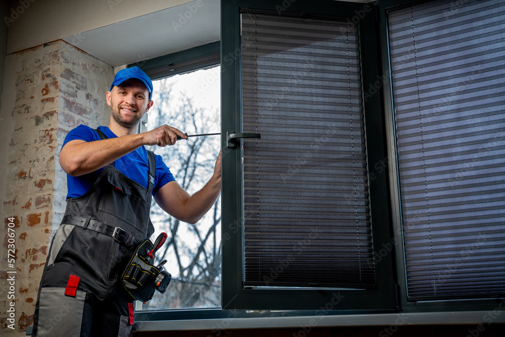 Builder in uniform repairs a plastic window with a screwdriver indoors. 