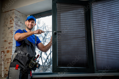 Builder in uniform repairs a plastic window with a screwdriver indoors. 
