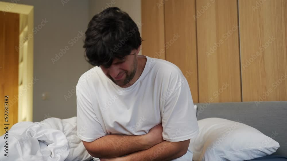 Man suffering from abdominal pain. A man in a white T-shirt is holding his stomach while sitting on the bed in the bedroom. Man suffering from abdominal pain. A man in a white T-shirt is holding his stomach while sitting on the bed in the bedroom.