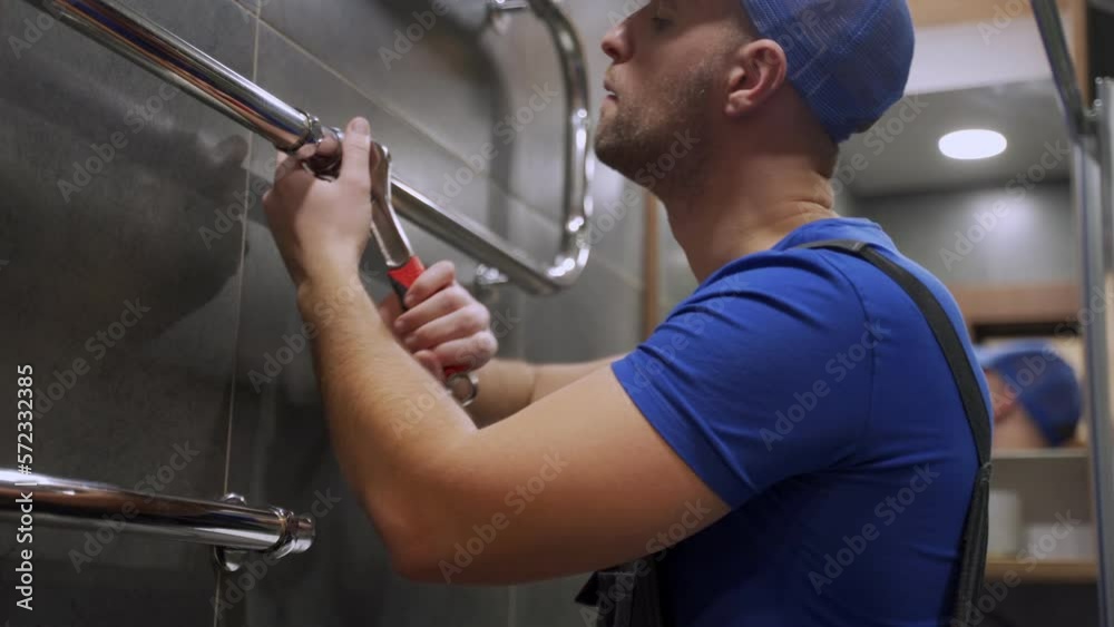 A plumber, a worker in uniform, is working, repairing a towel rail on the bathroom wall with a wrench. The concept of construction, maintenance and repair A plumber, a worker in uniform, is working, repairing a towel rail on the bathroom wall with a wrench. The concept of construction, maintenance and repair
