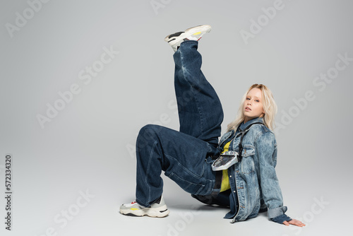 full length of young blonde model in denim outfit and trendy sneakers posing on grey.