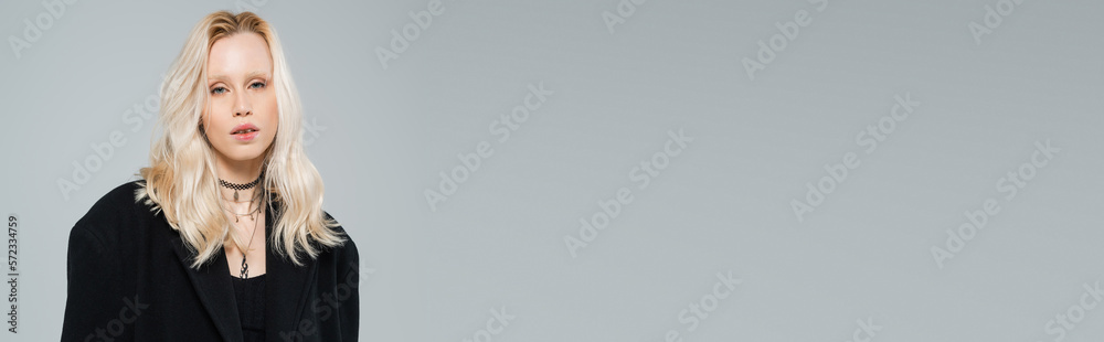 blonde and young woman in stylish black coat looking at camera while standing isolated on grey, banner. blonde and young woman in stylish black coat looking at camera while standing isolated on grey, banner.