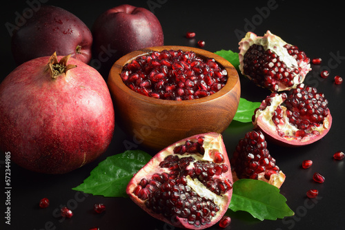 Wooden mortar full ripe arils of pomegranate, red apples, green leaves on black background. Still life fruits, sweet seeds. Split open clusters of pomegranates. Healthy food. Studio shot