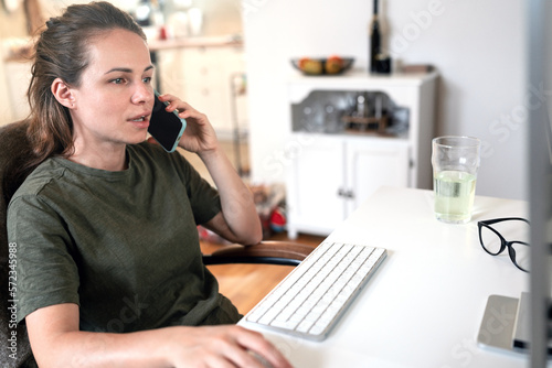 Young woman working from home working on computer and talking cell phone.