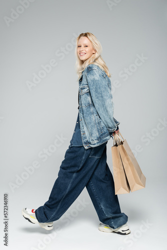 full length of happy young woman in stylish denim outfit holding shopping bags and walking on grey.