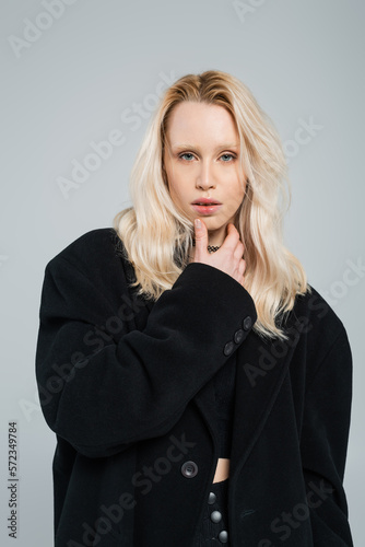 blonde and young woman in stylish black coat looking at camera isolated on grey.
