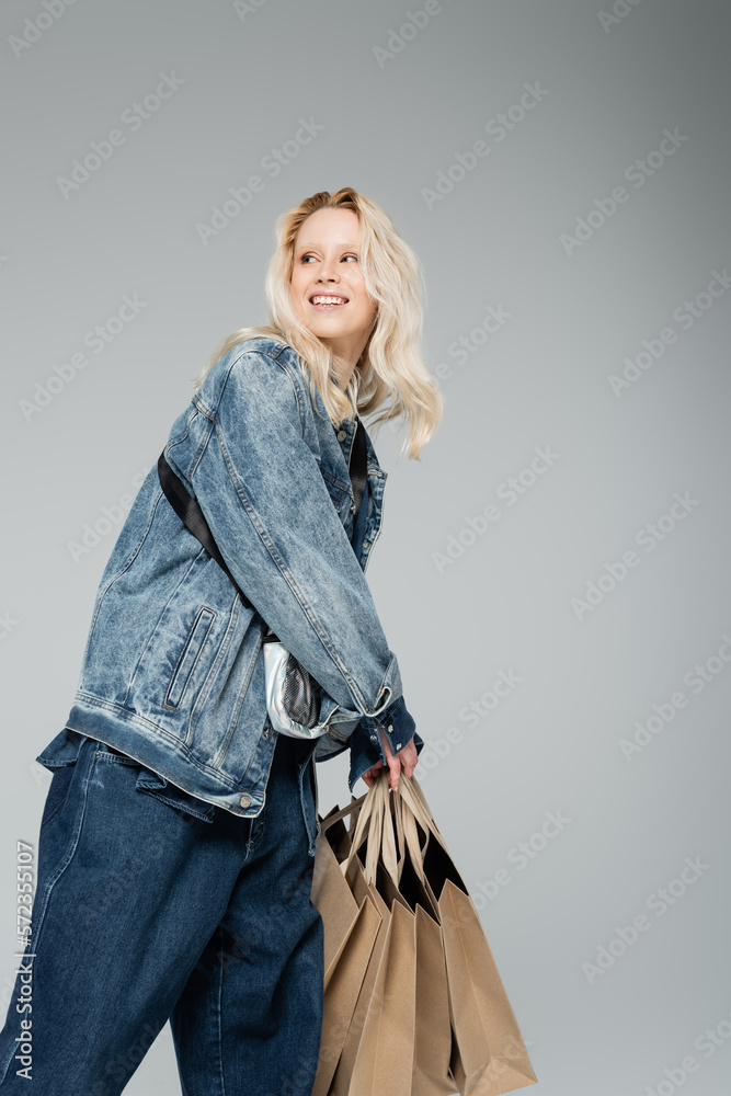 Fototapeta premium happy young woman in stylish denim outfit holding shopping bags and posing isolated on grey.