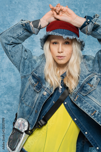 blonde woman in denim jacket and panama hat posing with raised hands near blue textured background.