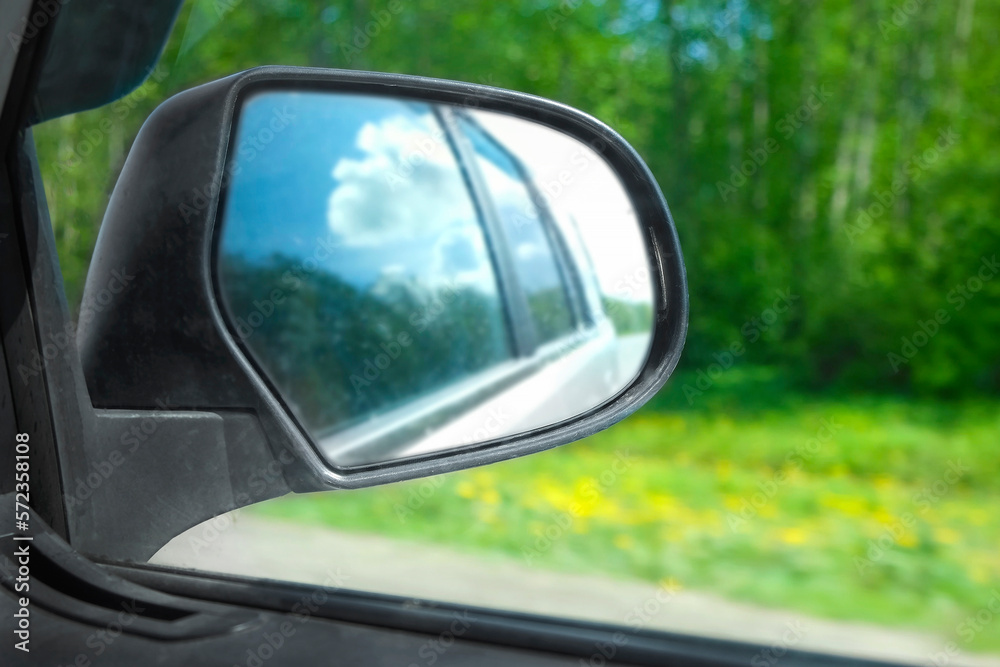 Side view mirror reflection of car auto in motion on road. Highway along green forest and trees in summer. Tourism and travel, journey trip concept. Beautiful nature and landscape. Car speeding road