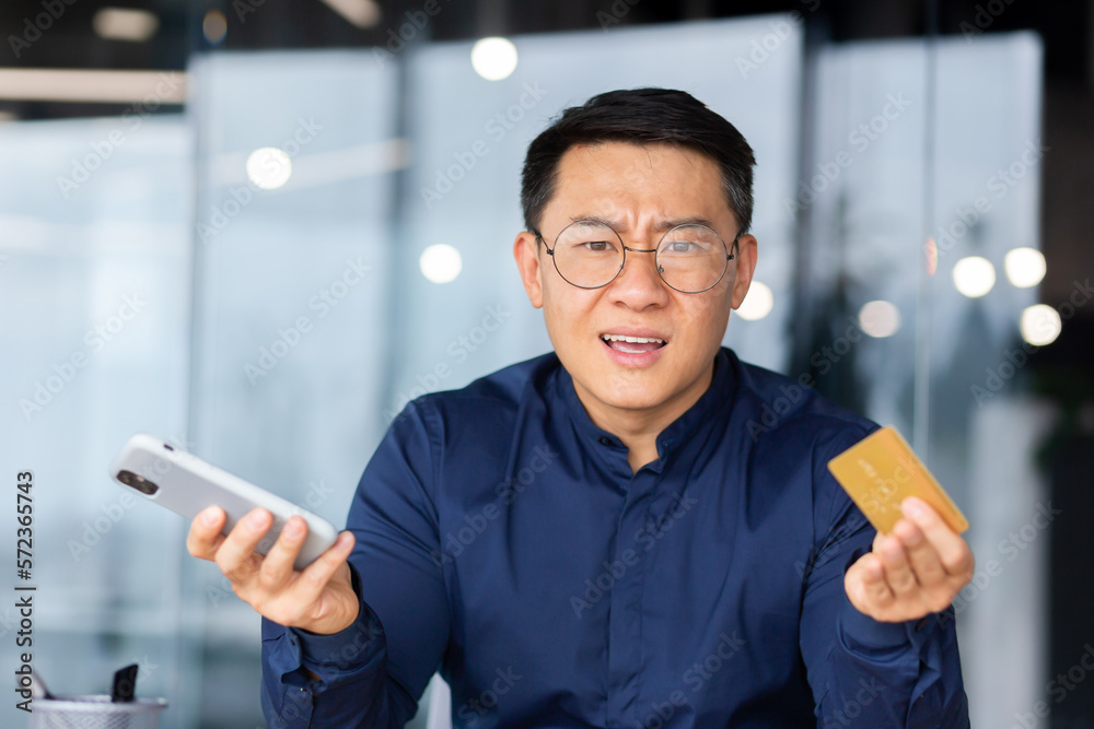Portrait of disappointed and angry businessman inside office, cheated asian man looking at camera upset holding bank credit card and phone, man got rejection and error in online money transfer. Portrait of disappointed and angry businessman inside office, cheated asian man looking at camera upset holding bank credit card and phone, man got rejection and error in online money transfer.