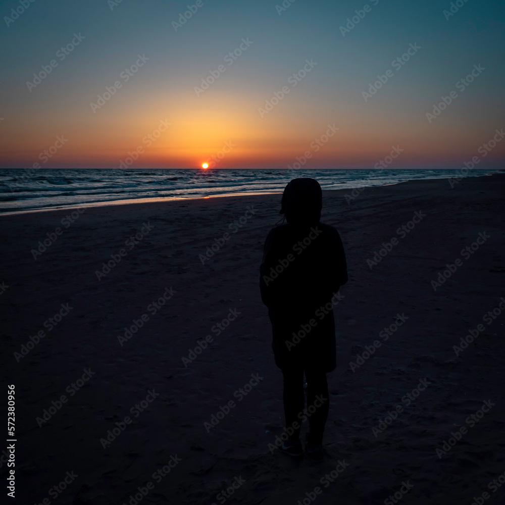 A young woman standing on a sea shore looking into a beautiful and colorful sunset on a calm as ice sea