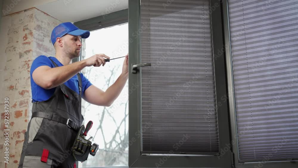 Builder in uniform repairs a plastic window with a screwdriver indoors. Builder in uniform repairs a plastic window with a screwdriver indoors.