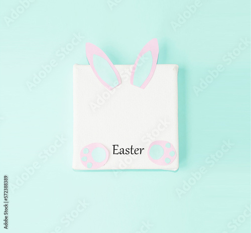 On a turquoise background, a white square background with rabbit ears and paws for writing text.  Concept holiday bright easter.  Copy space for text, flat lay, top view. © Yulia Shutikova