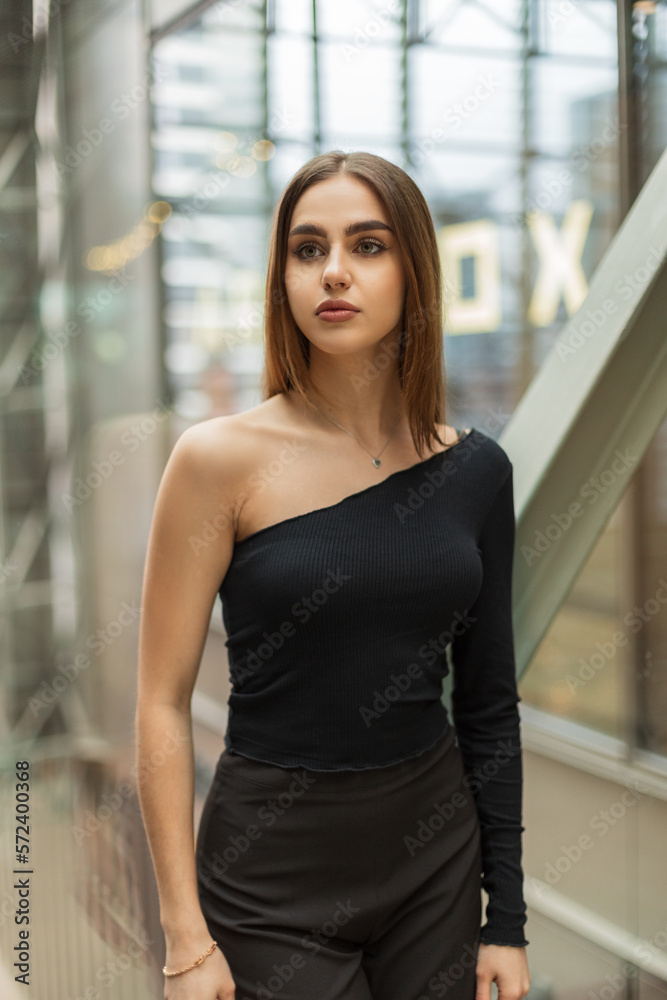 Stylish beautiful young girl model in fashionable black clothes with a one-shoulder top in an office building