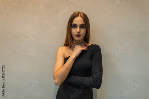 Female portrait of a beautiful fashion young girl in stylish black clothes with a one-shoulder top near a gray wall