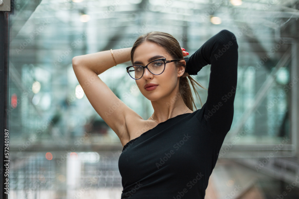 Stylish beautiful manager woman with glasses and a trendy black top stands and fixes her hair in a modern glass office building