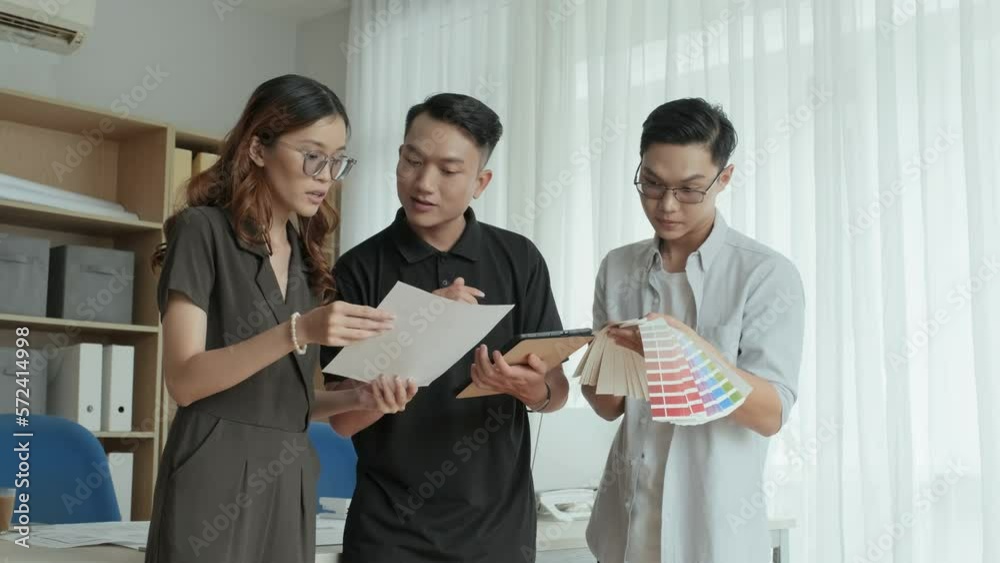 Medium shot of group of young Asian designers discussing paper plan and color palette while working together on project in office Medium shot of group of young Asian designers discussing paper plan and color palette while working together on project in office