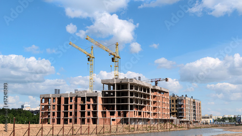 View of a construction site with multi-storey residential buildings under construction and several cranes against a blue sky with clouds.