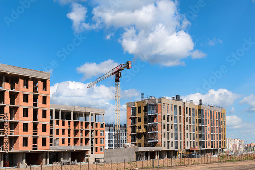 View of a construction site with multi-storey residential buildings under construction and a crane against a blue sky with clouds.