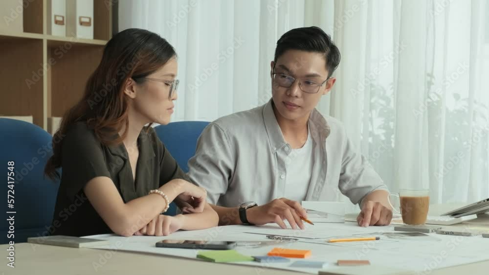 Young Asian students sitting at desk and discussing blueprints while working together on architectural project in study room Young Asian students sitting at desk and discussing blueprints while working together on architectural project in study room
