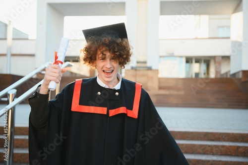 Graduation from university. Young smiling boy university graduate in traditional bonet and mantle standing and holding diploma in hand over university building background