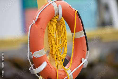 A vibrant orange polyethylene life ring hanging on a wooden post with long orange float line. There is reflective table on the preserver with white grab rope. The rescue equipment is a buoyant throw. 