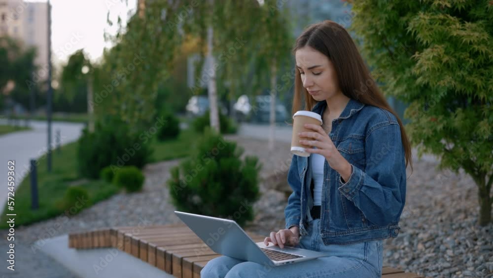 Caucasian Woman Sitting on the Bench Studying on the Laptop in the City Centre Drinking Coffee. Female Student Working Studying, Using Laptop for Studying Purposes. People and Technology Concept Caucasian Woman Sitting on the Bench Studying on the Laptop in the City Centre Drinking Coffee. Female Student Working Studying, Using Laptop for Studying Purposes. People and Technology Concept
