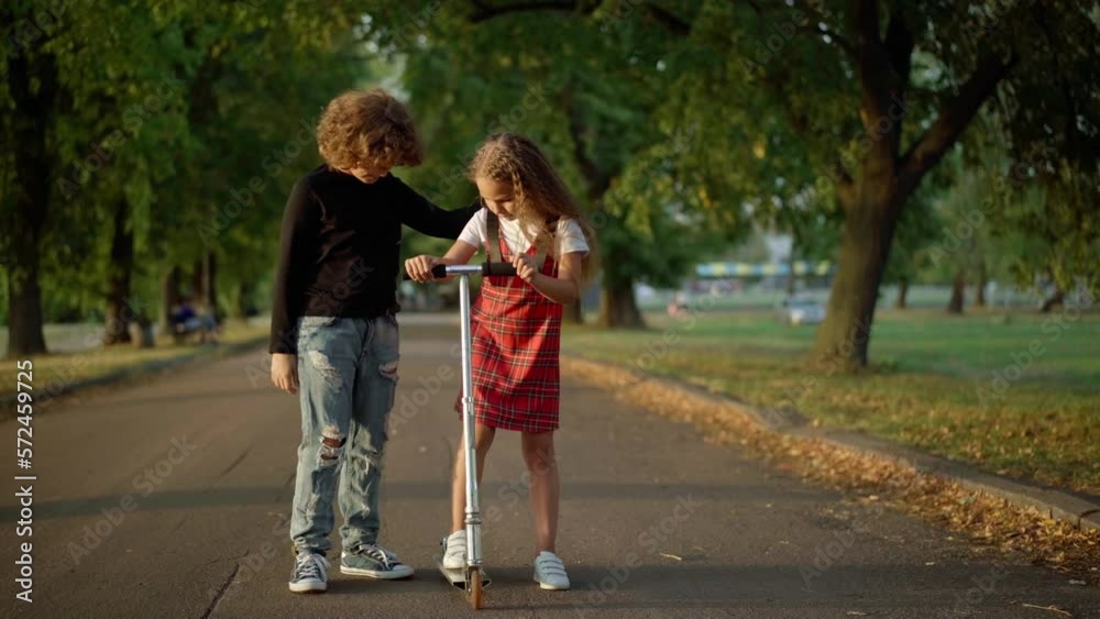 Wide shot Caucasian boy talking to girl teaching riding kick scooter. Curious child learning to ride with support of good friend Wide shot Caucasian boy talking to girl teaching riding kick scooter. Curious child learning to ride with support of good friend
