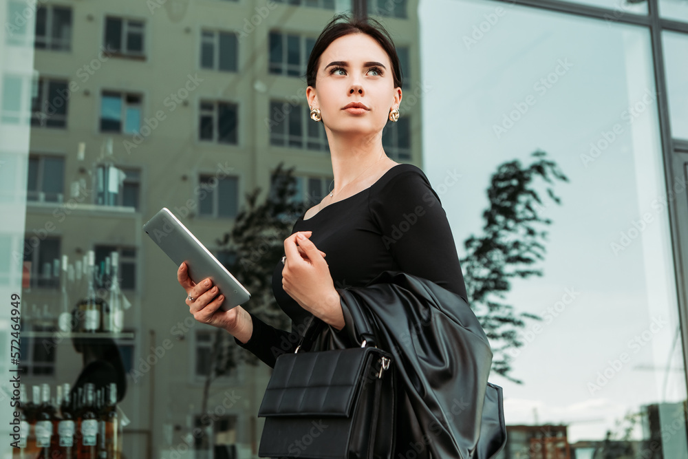 Fototapeta premium Pretty brunette caucasian woman dressed leather suit with shoulder bag walking outside hold tablet pc, order taxi, video chat call, typing message texting chatting, search information on the Internet 