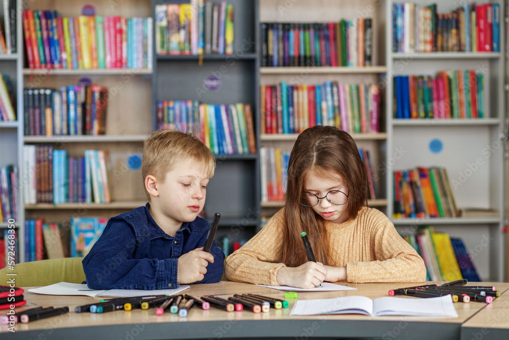Two schoolchildren write and draw sitting at table in library. Classmates study in classroom. Two schoolchildren write and draw sitting at table in library. Classmates study in classroom.