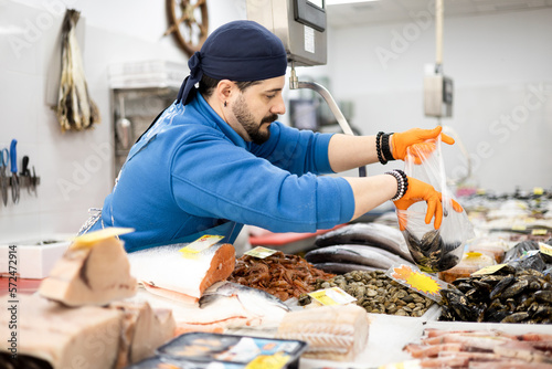 Foto A middle-aged fishmonger in an apron and hat is filling a plastic bag with mussels for a customer, small and medium business concept