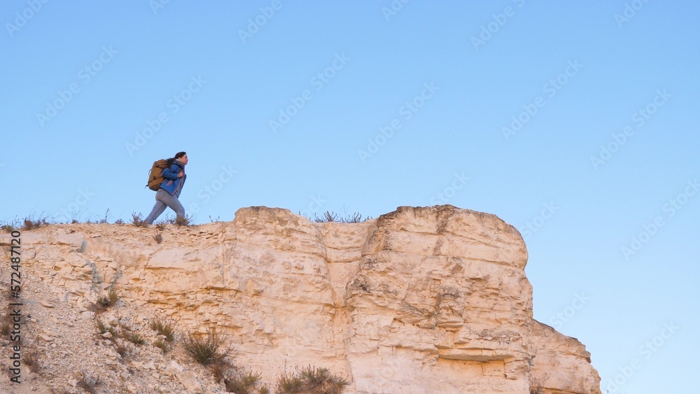 Girl hiker on top of cliff against background of blue sky. Happy woman traveler approaches edge of mountain with raised hands and enjoys beautiful landscape. Free woman tourist stands on edge of cliff