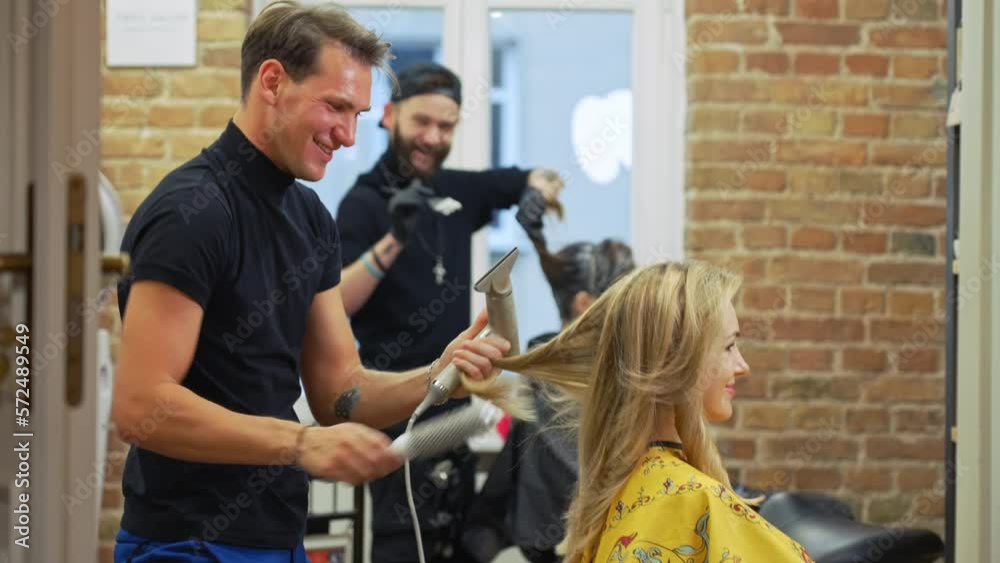 Two male hairdressers laughing at each other. They wave at each other. Customers sitting on chairs. Both clients are women. High quality 4k footage