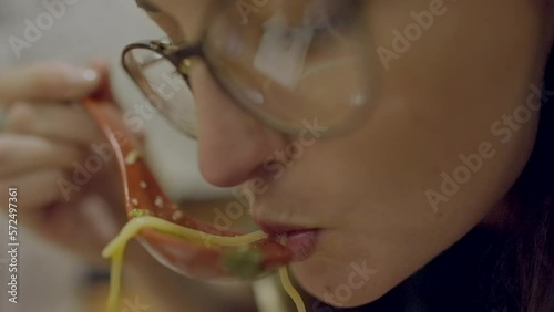This close up video shows a caucasian woman wearing eye glasses and slurping ramen from a spoon.
