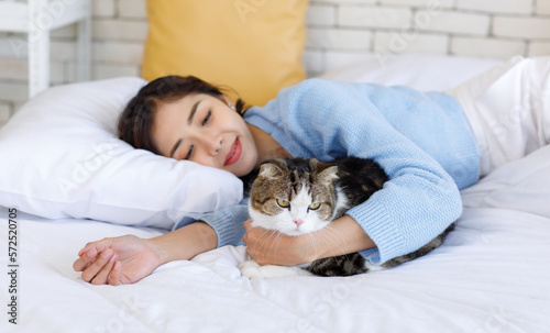 Millennial Asian young kindly cheerful female owner laying lying down on bed holding hugging cuddling showing love with short hair cute little domestic kitten furry pussycat pet friend on blanket