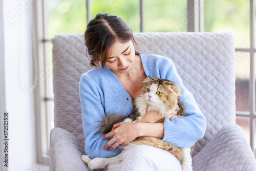 Millennial Asian young female owner sitting on cozy armchair smiling holding hugging cuddling looking showing love to cute fat tabby long hair little domestic cat furry purebred pussycat in arms