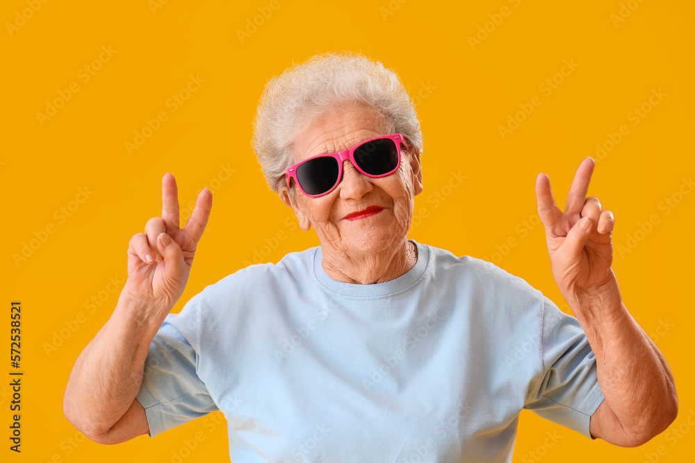 Naklejka premium Senior woman in sunglasses showing victory gesture on yellow background