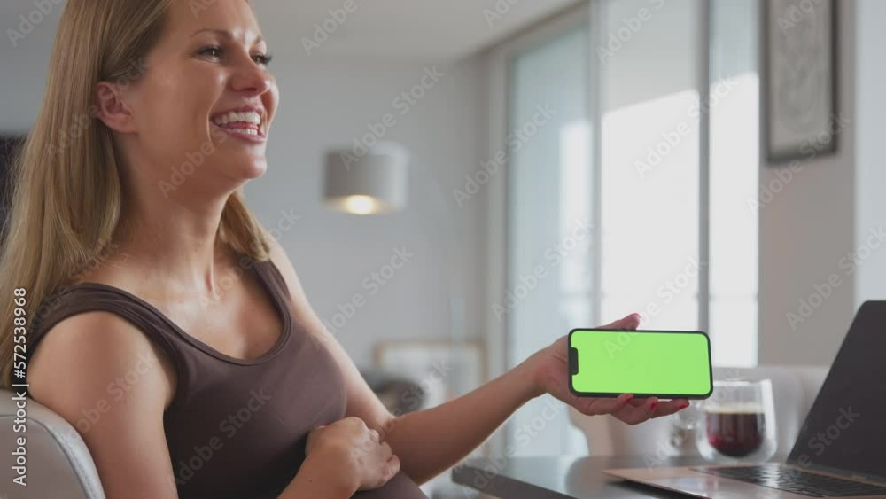 Smiling pregnant woman with hand on stomach at home showing green screen mobile phone to visiting off camera friend - shot in slow motion Smiling pregnant woman with hand on stomach at home showing green screen mobile phone to visiting off camera friend - shot in slow motion