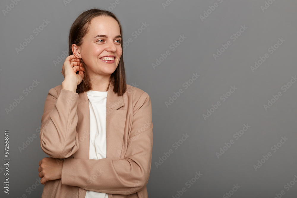 Image of attractive delighted woman with brown hair wearing beige jacket, smiling while looking aside at mockup for advertisement, posing isolated over gray background. Image of attractive delighted woman with brown hair wearing beige jacket, smiling while looking aside at mockup for advertisement, posing isolated over gray background.