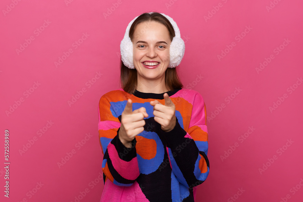 Indoor shot of smiling cheerful woman wearing sweater and earmuffs, pointing to camera, showing direction and choosing, indicating with index finger, posing isolated over pink background. Indoor shot of smiling cheerful woman wearing sweater and earmuffs, pointing to camera, showing direction and choosing, indicating with index finger, posing isolated over pink background.