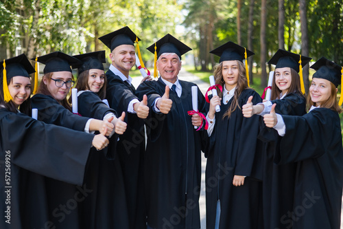 A group of graduates in robes give a thumbs up outdoors. Elderly student.