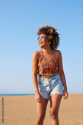 Laughing woman walking on beach. Portrait of laughing brunette with afro hairstyle in sunglasses and summer top and denim shorts looking sunward walking against clear blue sky on sunny day.