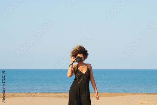 Woman in black dress with camera against the sea. Portrait of curly haired woman in black dress taking photo of photographer with digital camera. She is standing in the wind against blue sea and sky