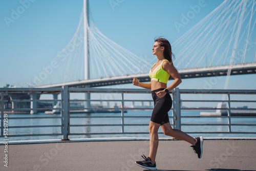Fotografía Young brunette woman in sportswear running at embankment  against huge bridge on background