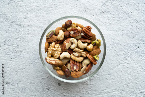Viewed from above, against a textured concrete background, an isolated glass bowl holds a large variety of shelled nuts.