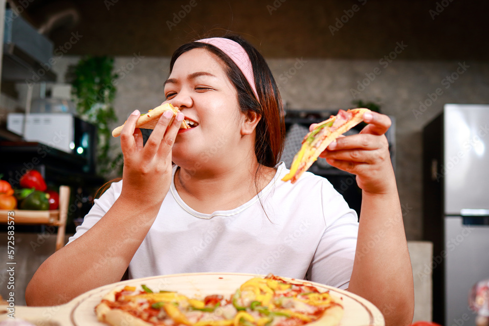 Fat Asian happy woman eating pizza, she holds two slices of pizza and eats deliciously in the kitchen at home. Fast food concept. weight loss Fat Asian happy woman eating pizza, she holds two slices of pizza and eats deliciously in the kitchen at home. Fast food concept. weight loss