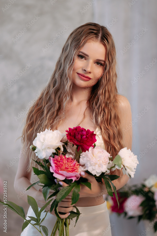 Attractive woman with long blond wavy hair stands with a bouquet of flowers in her hands in a bright interior. The concept of femininity and congratulations on International Women's Day.